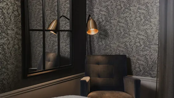 Ett mysigt hotellrumshörn med mönstrad tapet, en bekväm fåtölj och en varm lampa som reflekteras i en spegel.
