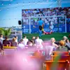 Folk samlet udendørs, ser en fodboldkamp på en stor skærm med lyskæder over. Resultatet er 27:21 SLO 0 0 SER.