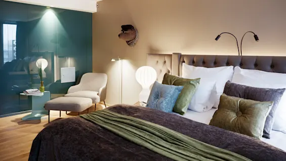 A bed with green pillows and a blanket sits against a beige wall. Nearby, there's a chair with an ottoman, a green side table, and decorative lighting.