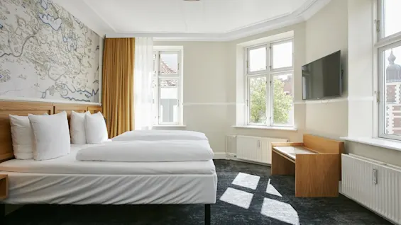 Et lyst hotelværelse med to senge, korttapet og solbeskinnede vinduer.
