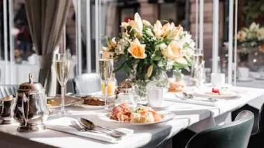 Et pent dekket frokostbord med champagne, bestikk og en fargerik blomsteroppsats i et solfylt rom med vinduer på Hotel Kämp i Helsinki.