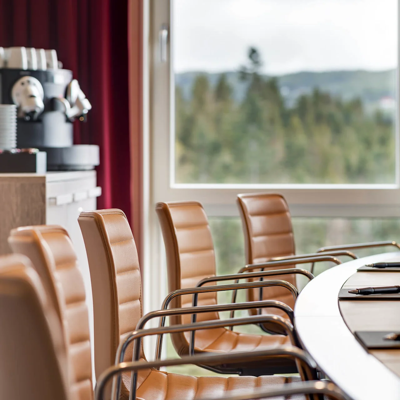 Konferansestoler står rundt et bord med notatblokker og penner. En kaffemaskin med stablede kopper er nær et vindu som viser trær.