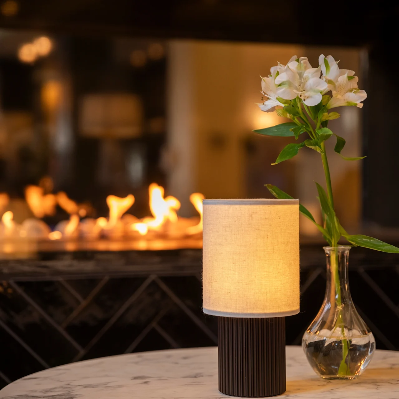A table lamp and vase with flowers sit on a marble table, with a fireplace softly illuminating the background.