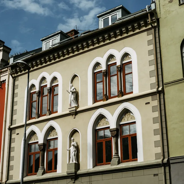 Udsmykkede bygningsfacader med buede vinduer og dekorative statuer under en blå himmel. En blanding af arkitektoniske stilarter og farver.