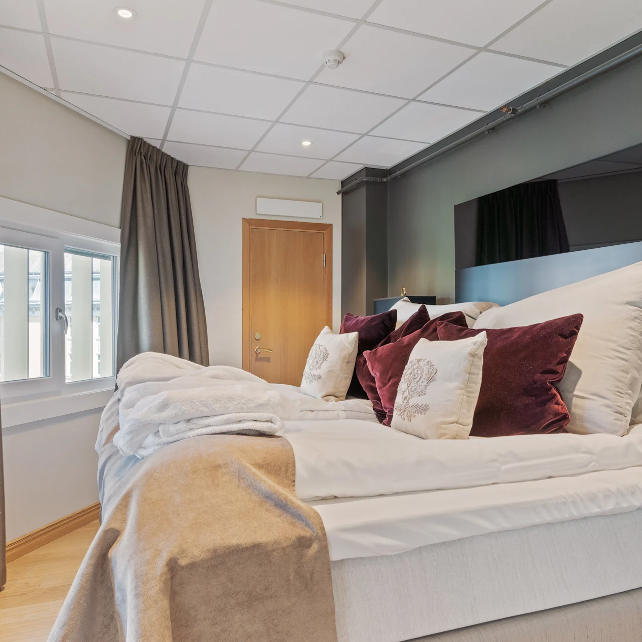 A bed with white linens and burgundy pillows is unmade in a modern bedroom. Thick curtains adorn the windows, and a wooden door is closed, enhancing the cozy atmosphere.