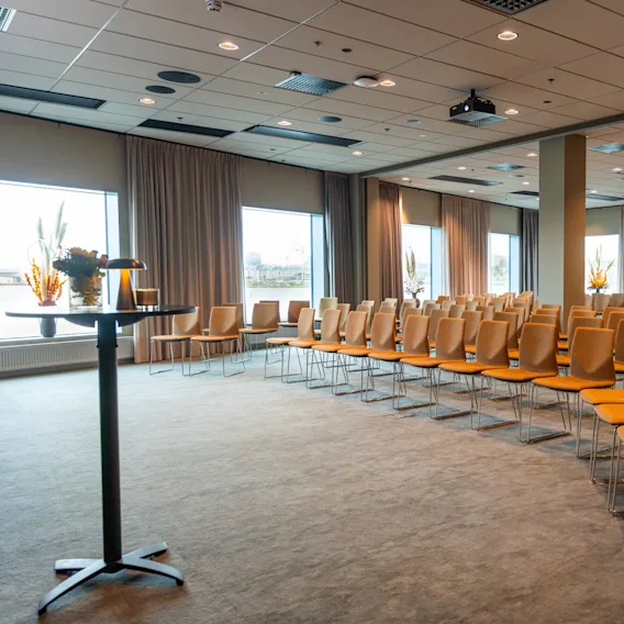 Rader med oransje polstrede stoler er plassert i et konferanserom. Store vinduer med gardiner og potteplanter skaper en åpen, luftig atmosfære.