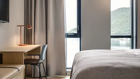 Et veloplyst hotelværelse med skrivebord, stol og lampe ved vinduet. Gardiner dækker delvist vinduet og afslører udsigt til et bjerg og vand udenfor.