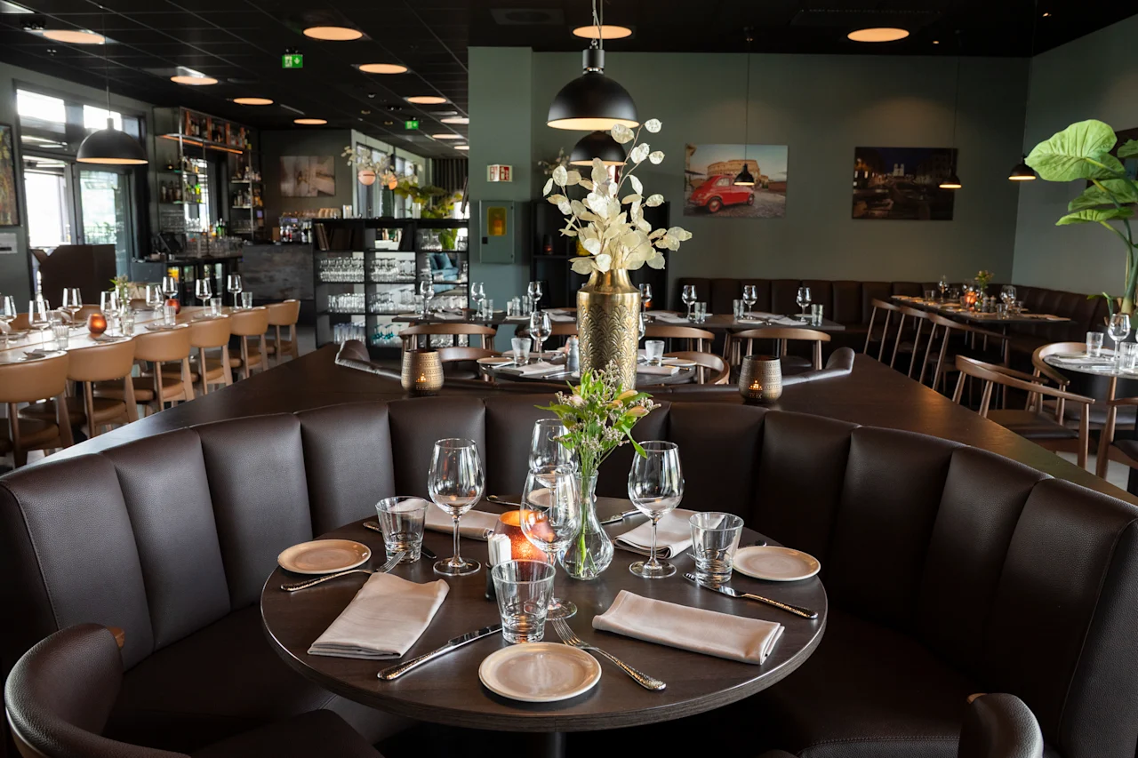 Et koselig restaurantinteriør med mørkebrune båser og bord dekket for bespisning, med et barområde i bakgrunnen.
