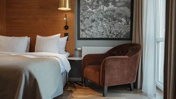 Et moderne hotelværelse med en komfortabel seng, en blød lænestol og et stort naturinspireret kunstværk, der skaber en afslappende atmosfære.