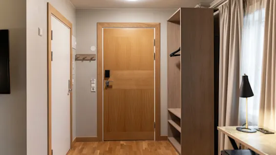 En innbydende hotellromsinngang med tredør, knagger, en åpen garderobe og et skrivebord med lampe ved vinduet.