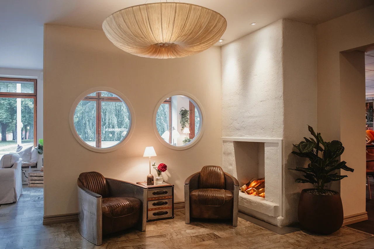 A cozy hotel lobby seating area with two leather armchairs, a glowing fireplace, and round windows looking out to trees.