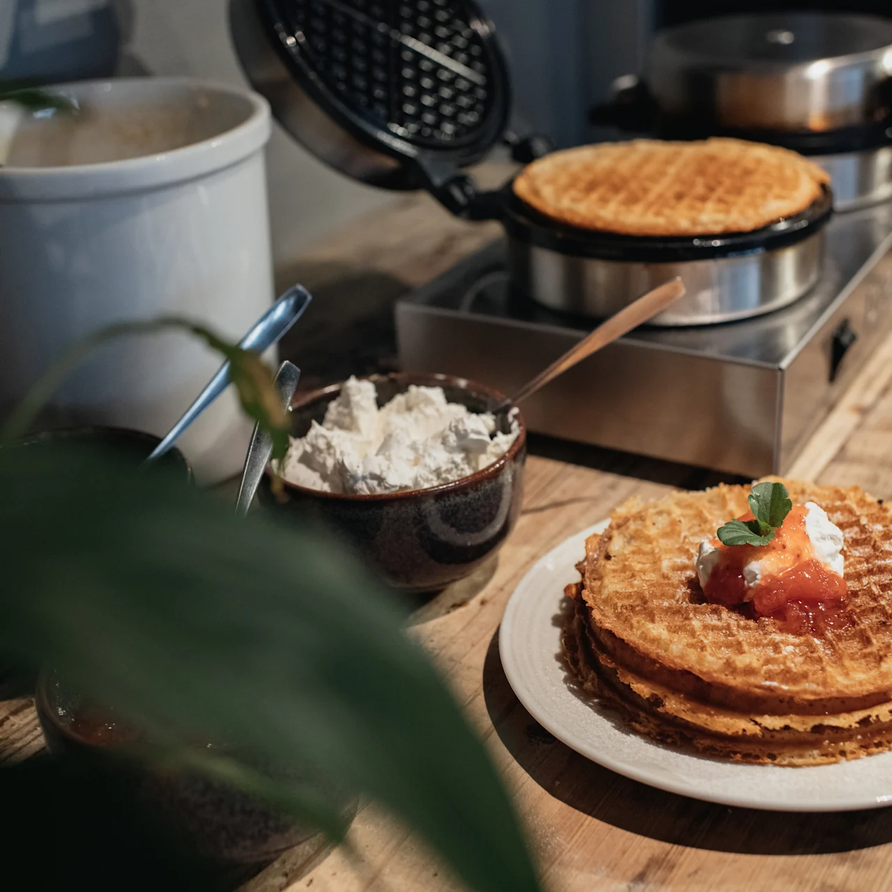 Waffles sit on a white plate topped with cream and fruit, surrounded by a waffle maker and bowls of additional ingredients on a wooden counter.