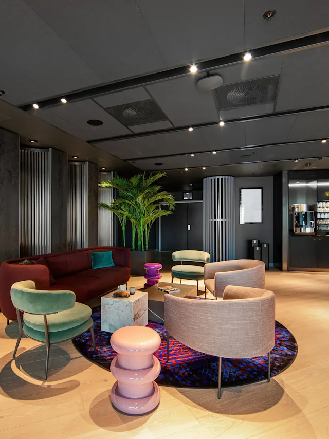 A modern lounge features stylish chairs, a burgundy sofa, and a circular rug. A potted plant decorates the space, and a beverage station is visible in the background.