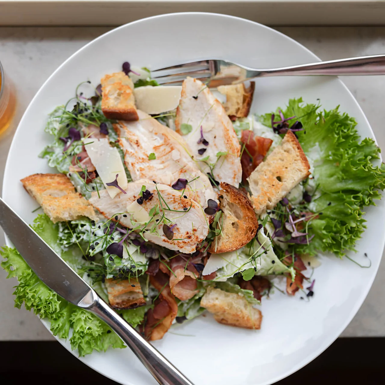 En tallerken med Caesar-salat med grillet kylling, croutoner, parmesanskiver og brøndkarse står på et bord ved siden af et glas med ravfarvet drik. En gaffel og kniv ligger på tallerkenen.