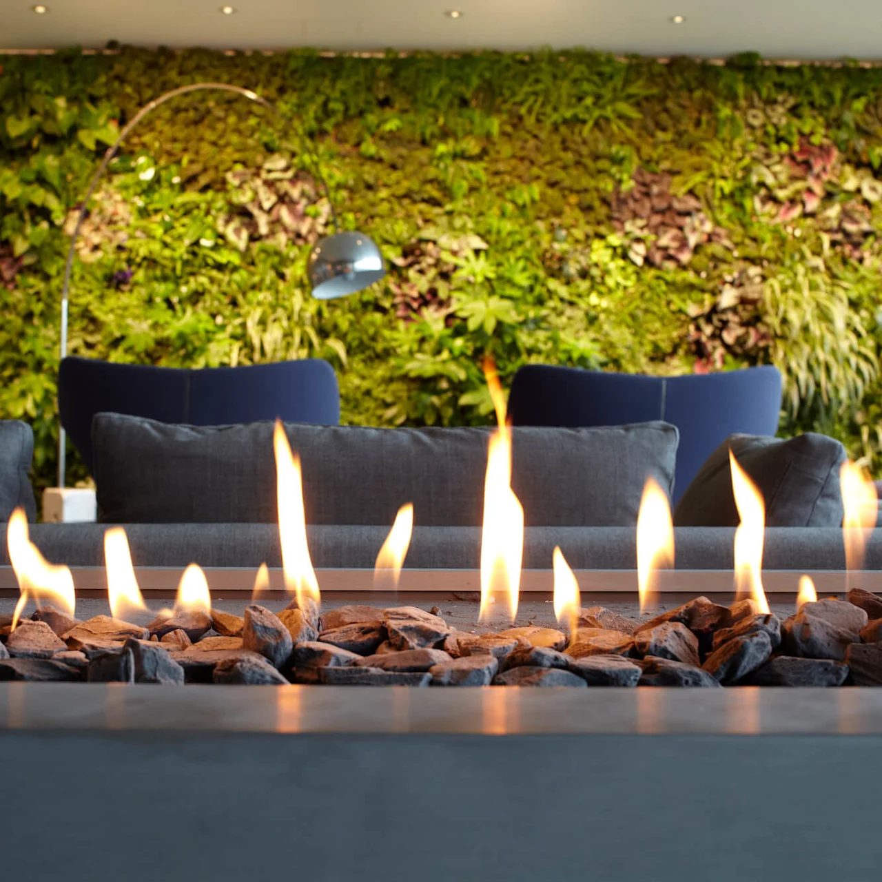 Fireplace with burning flames, surrounded by stones, placed in front of blue sofas. A lush green vertical garden wall is in the background, creating a cozy, modern atmosphere.