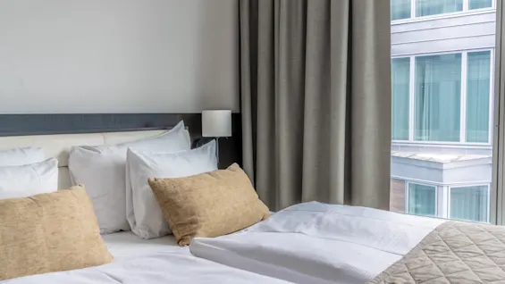 A hotel room with a comfortable bed, white linens, and brown pillows, offering a view of a modern building through the window.