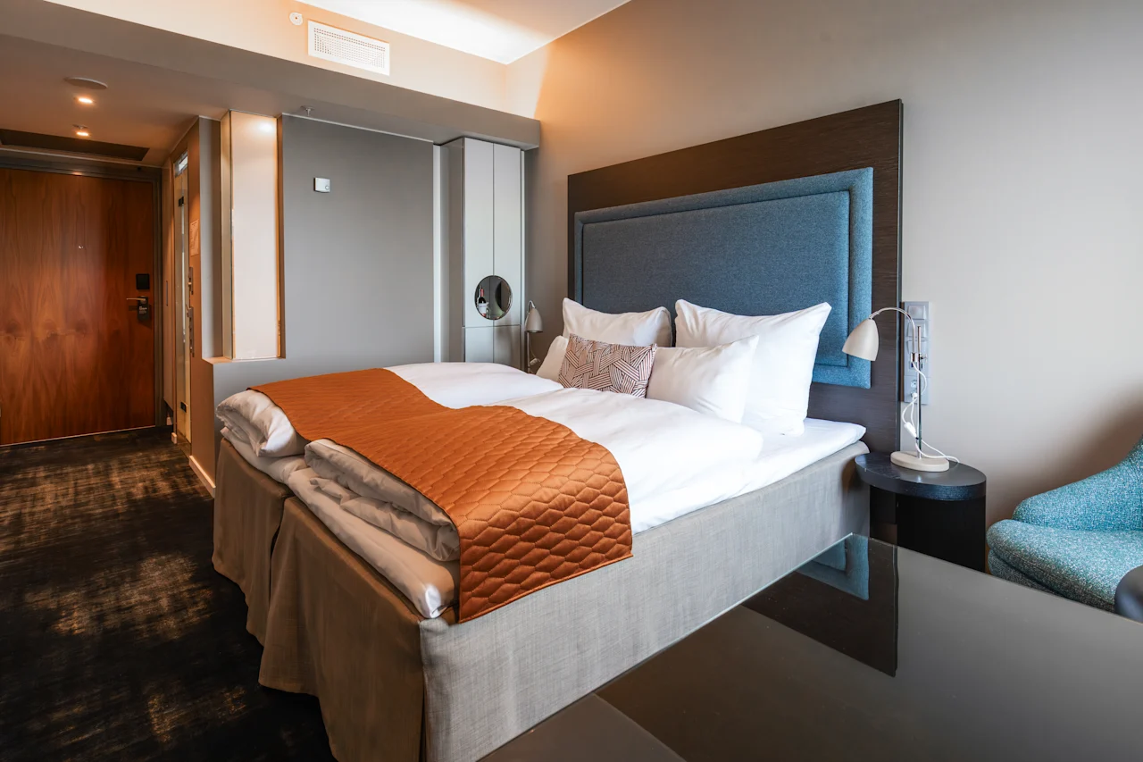 Siististi pedattu sänky, jossa on valkoinen vuodevaatteet ja oranssi peitto, sijaitsee modernissa hotellihuoneessa. Lähellä turkoosi tuoli ja tumma puuovi täydentävät neutraalia sisustusta.