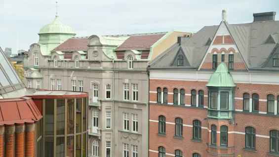 Rød og beige historisk bygning med udsmykkede facader og grønne kupler står ved siden af hinanden, set fra en hævet vinkel under en overskyet himmel.
