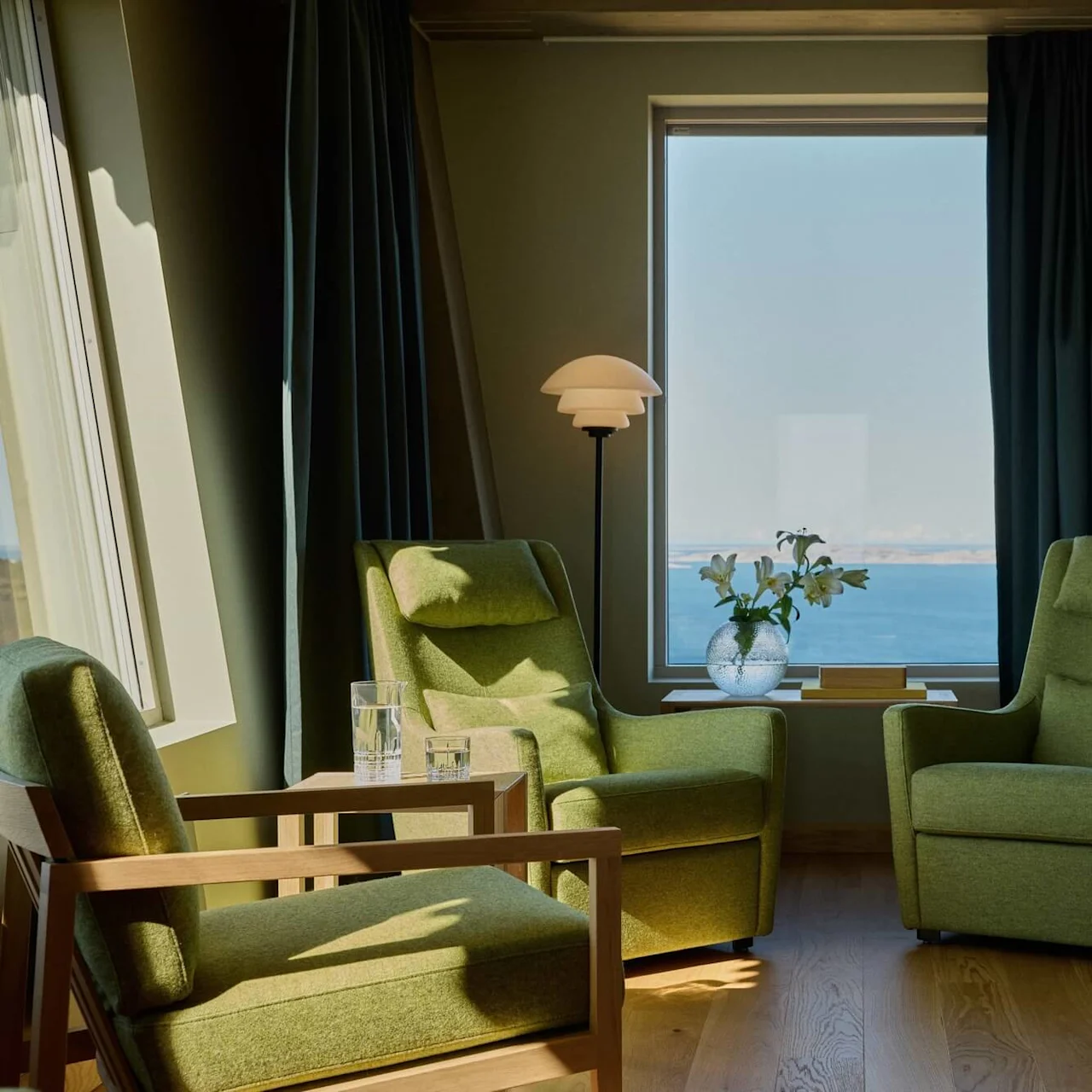 Green armchairs face each other beside a wooden table with glasses, a vase of flowers, and a lamp. They overlook a large window showing a sea view.