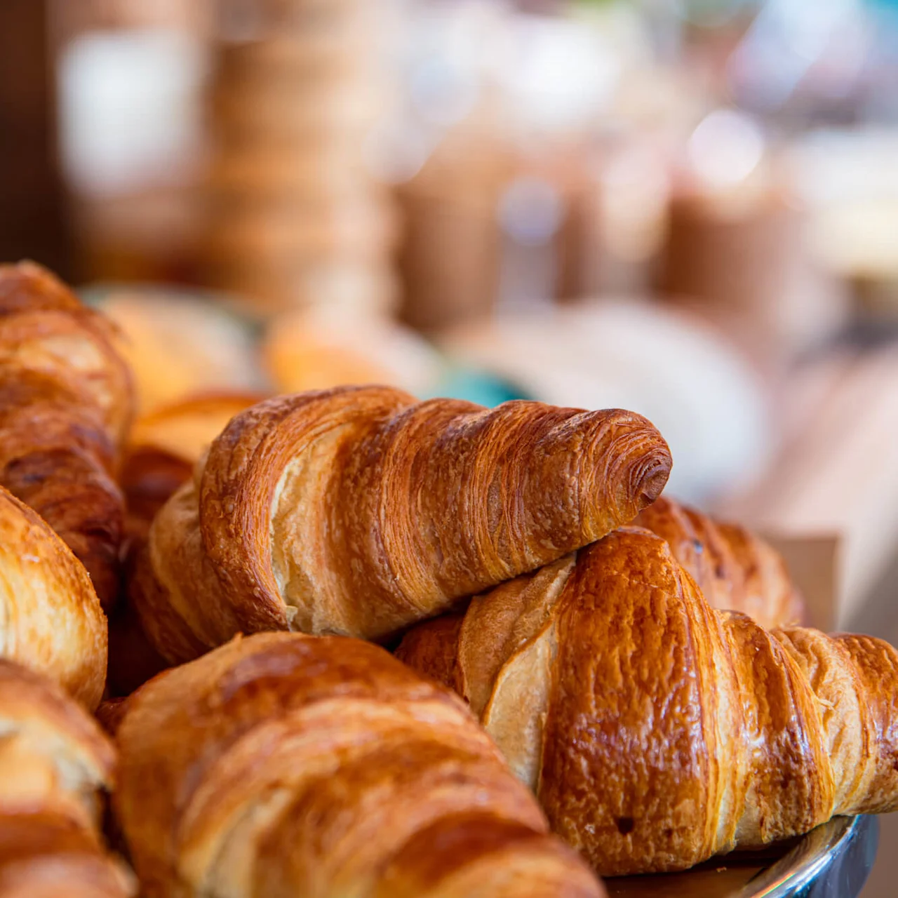 Golden croissants piled in a bowl, foreground focus, blurry café backdrop.