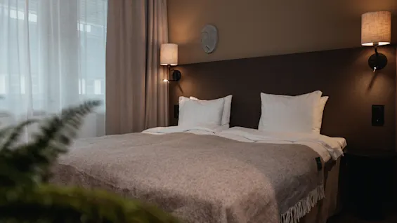 A neatly made bed with white linens and a gray blanket, flanked by two wall-mounted lamps, is in a softly-lit bedroom with curtains and a visible plant in the foreground.
