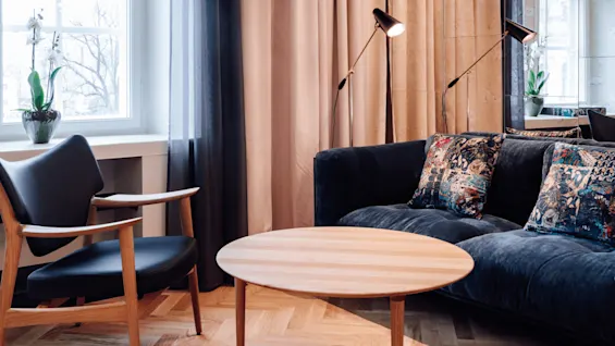 A wooden table surrounded by a black chair and blue sofa, adorned with patterned cushions. Two floor lamps illuminate the room with curtains and potted plants by a window.