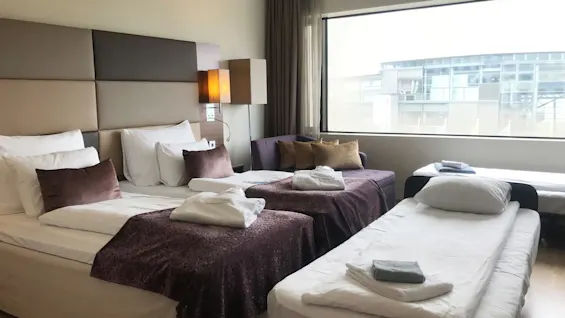 Two beds and a sofa sit in a hotel room, covered with white sheets and purple blankets. Robes and towels are neatly placed, with a large window offering an outside view.