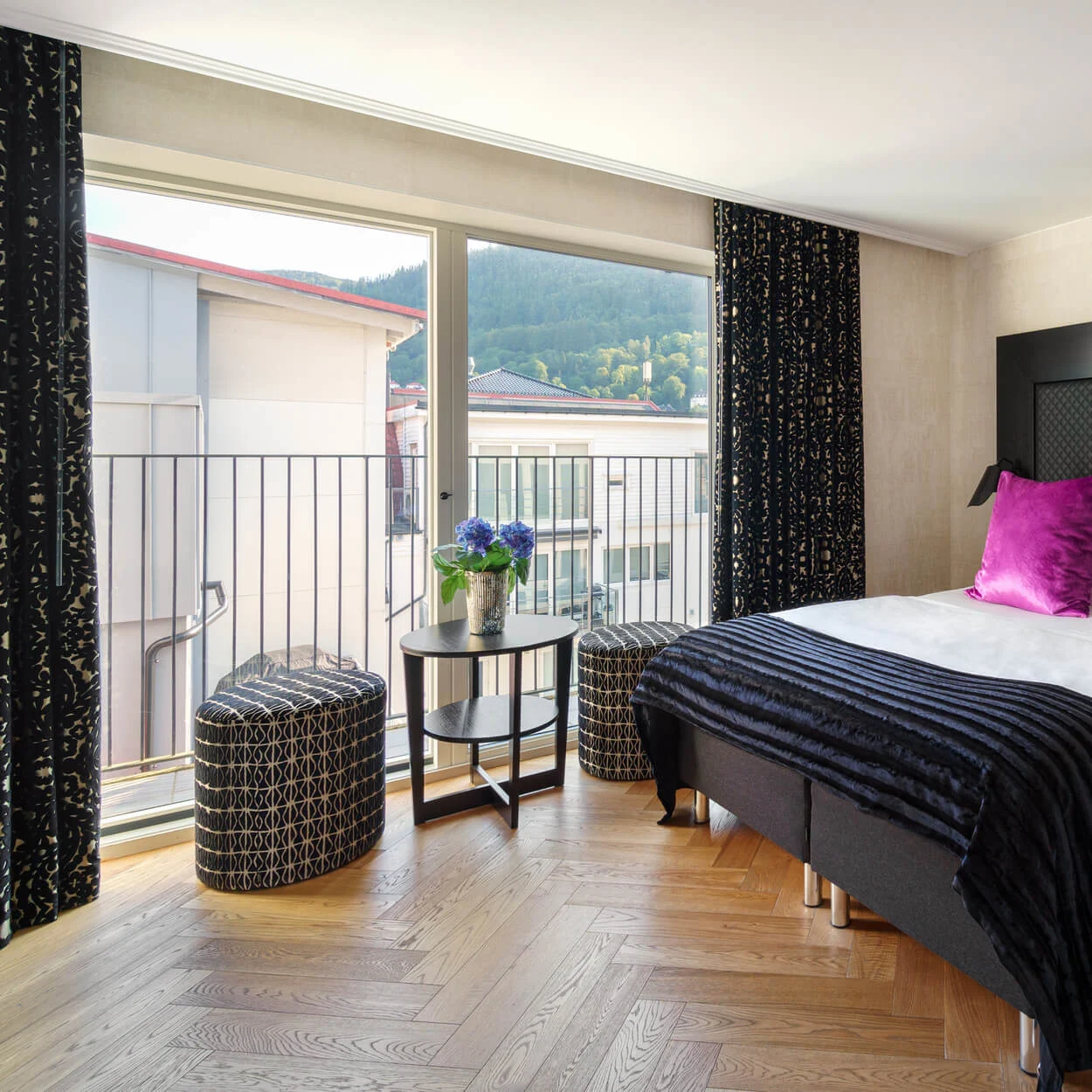 A bed with vibrant purple pillows is set in a modern room with wooden floors, beside a window overlooking buildings and greenery. A small table with flowers stands nearby.