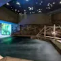 En rolig indendørs pool med let damp, omgivet af stenvægge. Et metalgelænder fører ned i vandet, under et loft med stjernelignende lys.