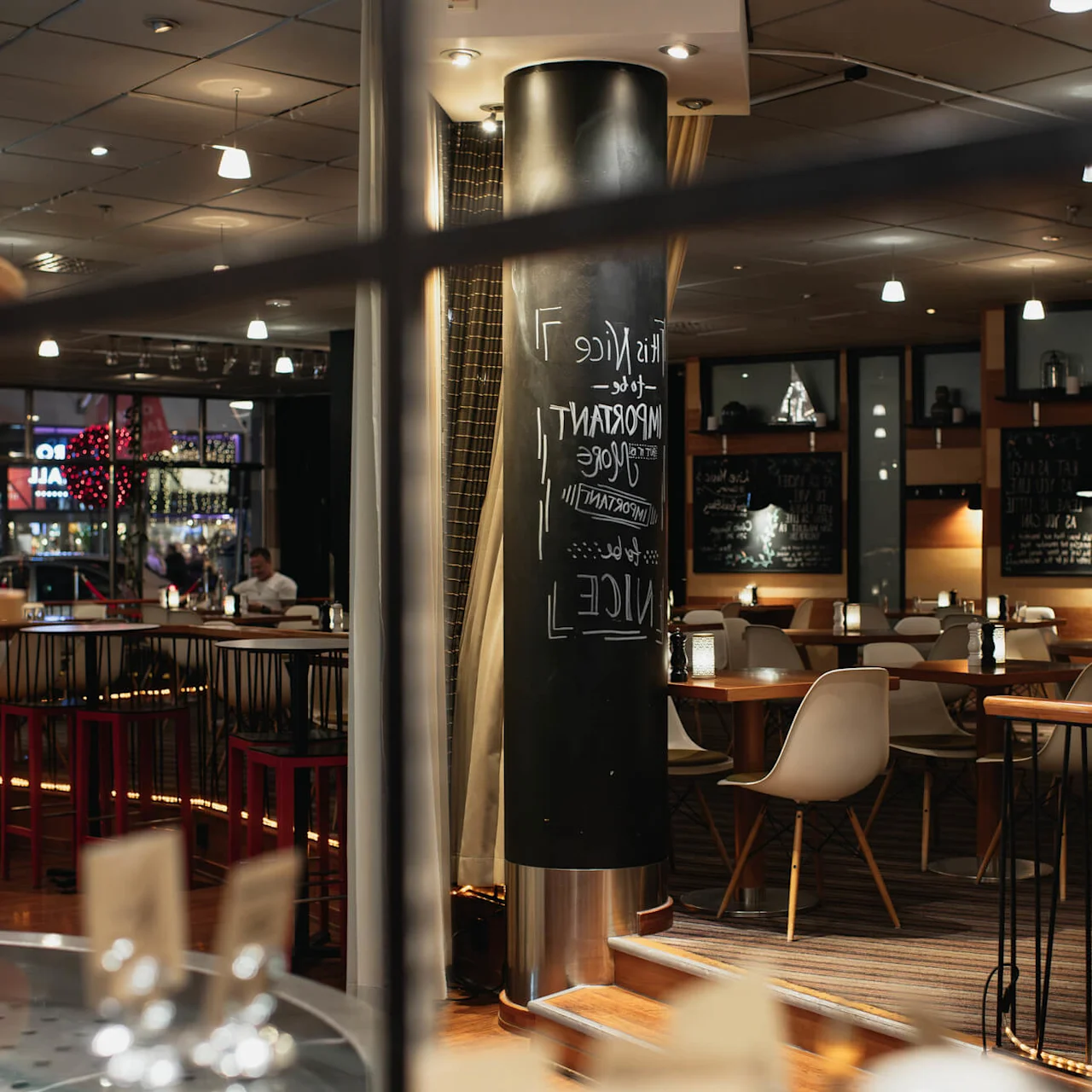 A dimly lit restaurant interior features empty tables and chairs. A prominent black column displays the text: "THIS NICE IMPORTANT LOVE". Through the window, colorful lights and people are visible outside.