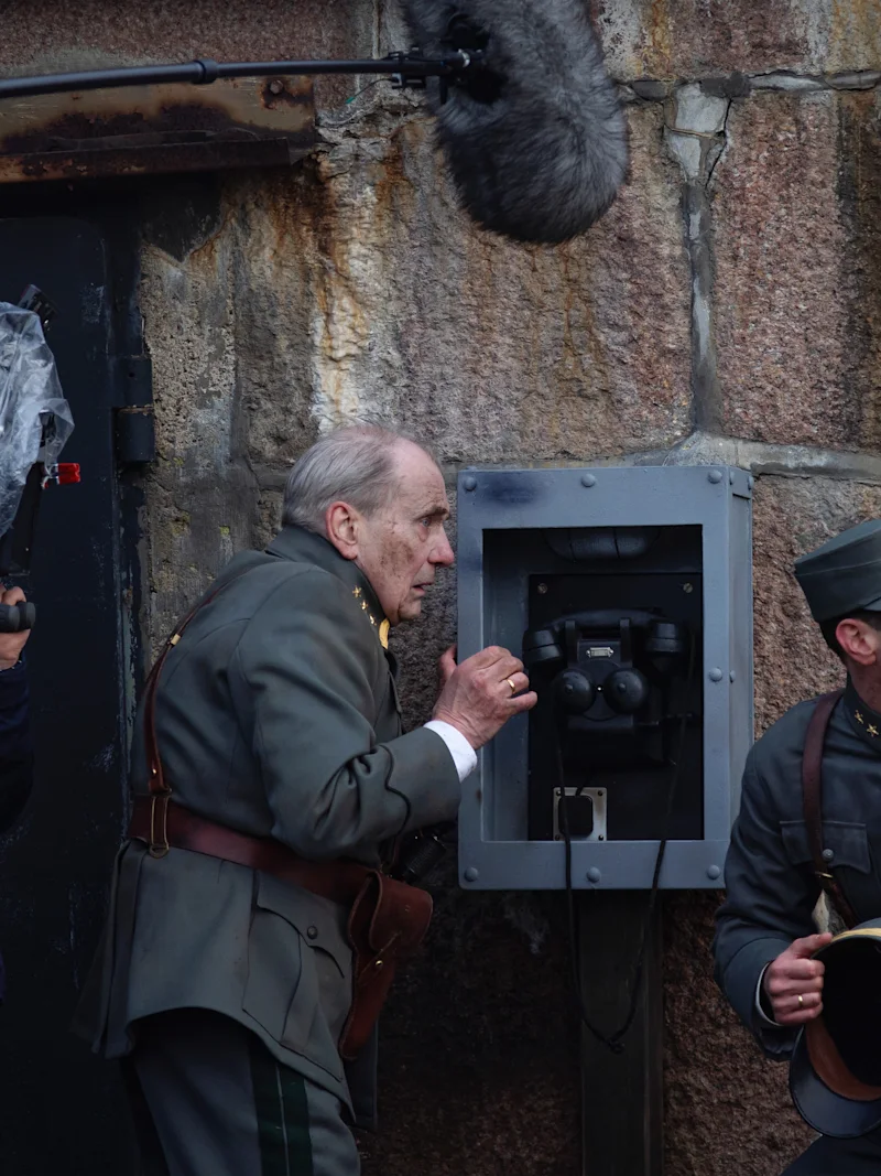 En kameraman filmar två skådespelare klädda som soldater som använder en gammal telefon på en vägg av sten. En skådespelare lyssnar uppmärksamt medan den andra hukar med en hatt i handen.