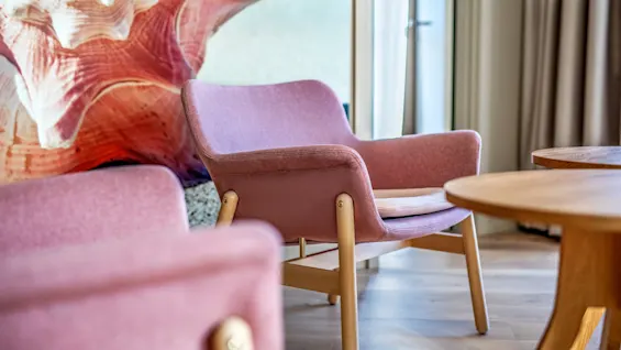 A pink armchair with wooden legs sits on light wooden flooring, flanked by a table. A large seashell mural decorates the wall, and a curtain-covered window brightens the room.