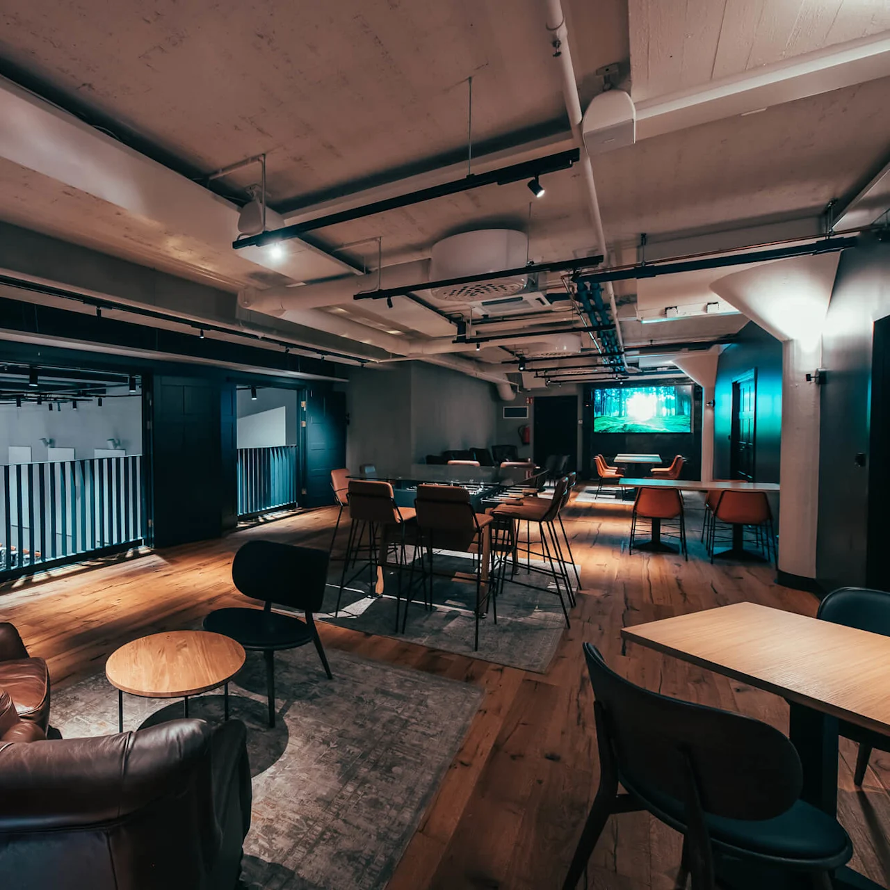 En romslig lounge med skinnstoler og runde bord, omgitt av moderne dekor og myk belysning, med en stor TV-skjerm mot veggen.