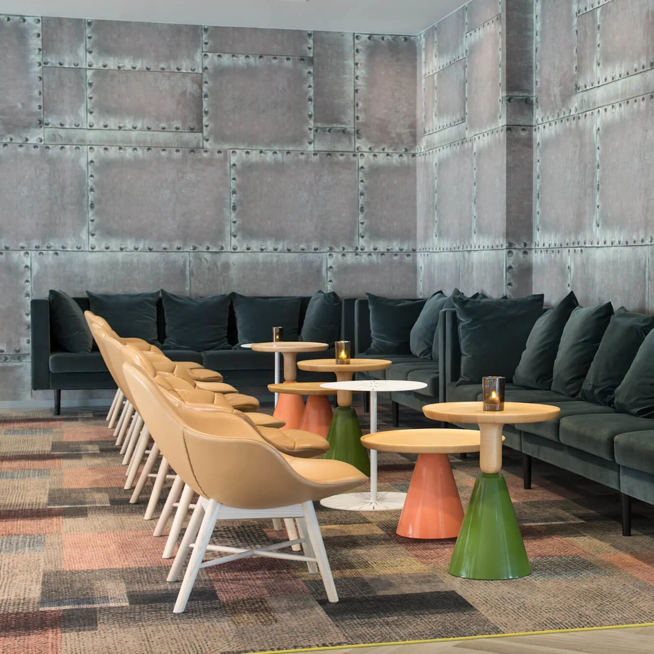 Dark green sofas with cushions line a textured, industrial-style wall. Beige chairs face colorful pedestal tables with candles, situated on a patterned carpet.