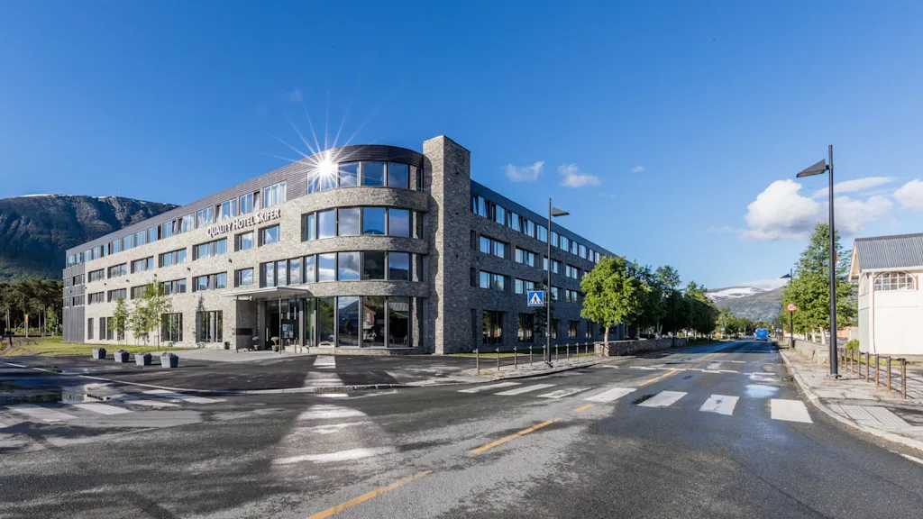 En moderne fleretagers bygning med navnet "Scandic Valdres Hotel" står på et gadehjørne, med reflekterende vinduer omgivet af træer, bjerge og en klar blå himmel.