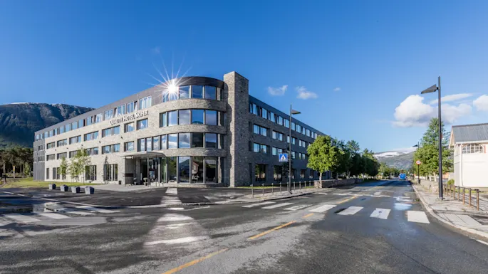 A modern, multi-story building labeled "Scandic Valdres Hotel" stands on a street corner, with reflective windows and surrounded by trees, mountains, and a clear blue sky.