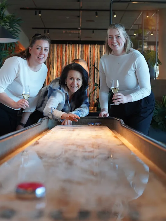 Tre personer spelar shuffleboard i en avslappnad miljö, ler och håller i drycker.