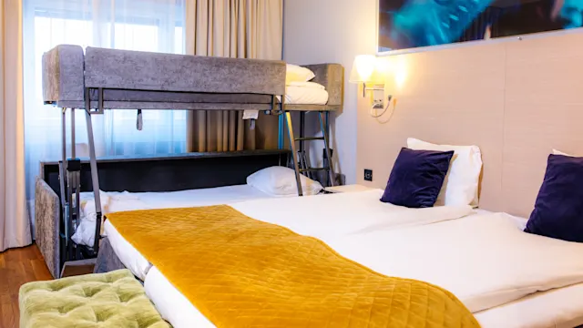 Ett hotellrum innehåller en stor säng med två kuddar och en orange filt, bredvid en våningssäng. Miljön är ljus, med ett fönster och beige gardiner.