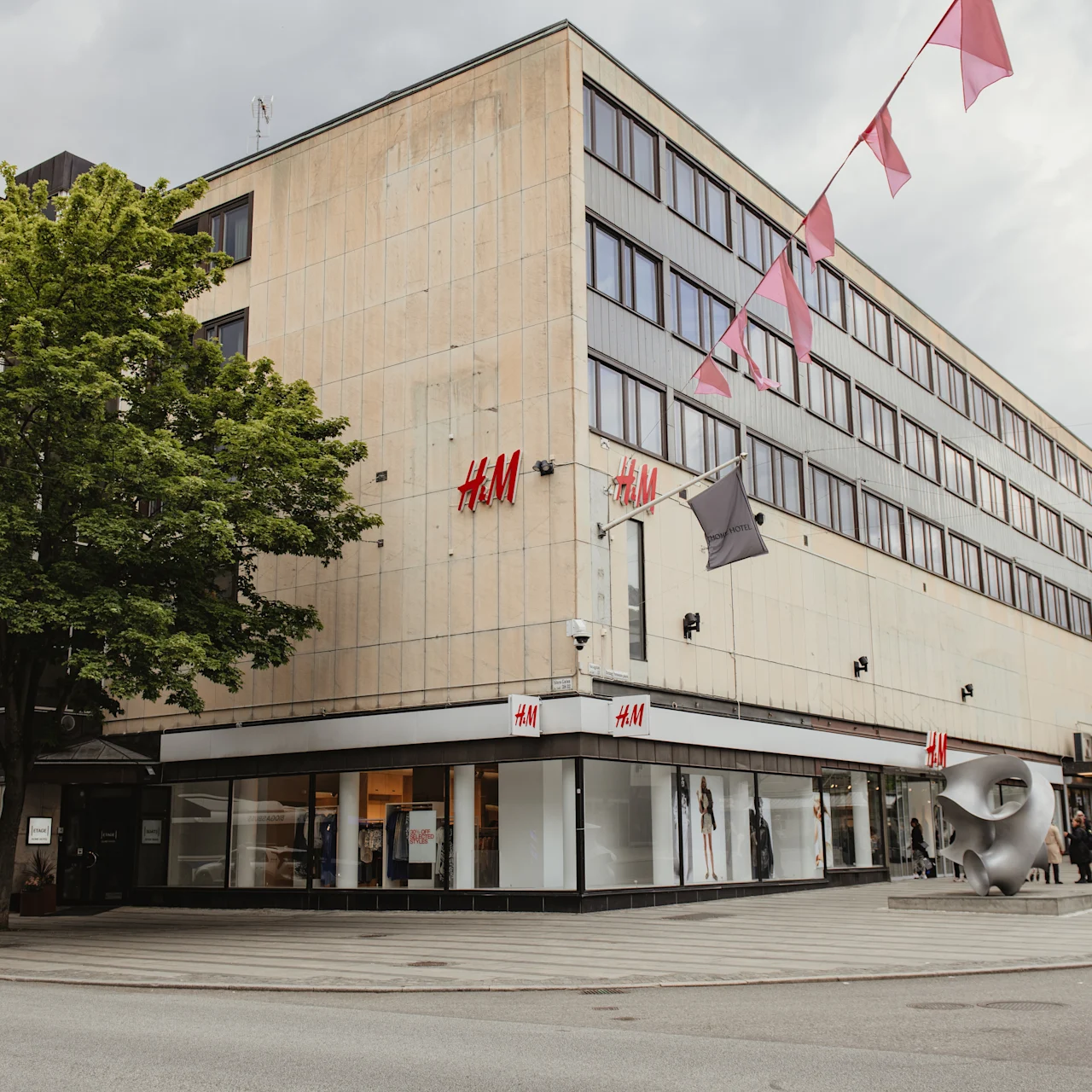 A corner building houses an H&M store with a tree in front. Colored flags hang overhead. People walk along the street. 