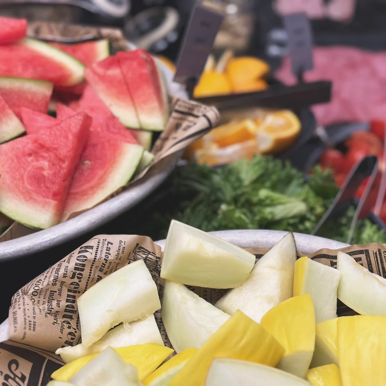 Watermelon and mixed vegetables lie in bowls on a table, surrounded by sliced cucumbers, tomatoes, and greens. Background has more sliced fruits and meats. Newspaper lining is visible.