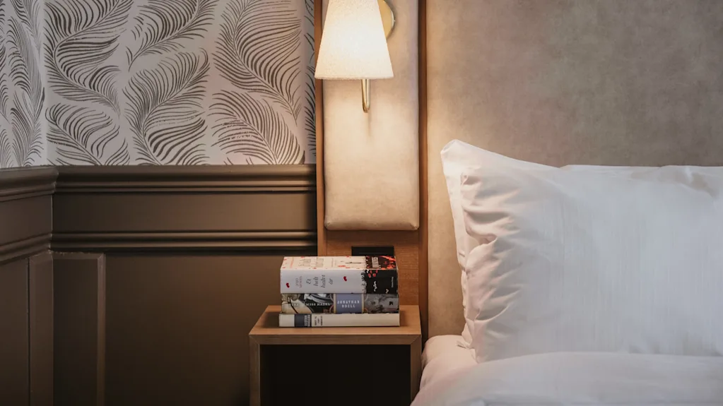 En hyggelig sengebordsscene med en stak bøger på et natbord, oplyst af en varm væglampe, ved siden af en komfortabel seng.