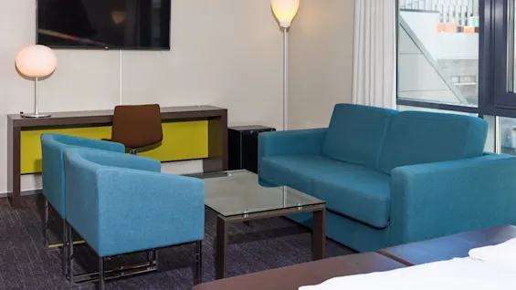 A blue sofa set stands in a modern hotel room, facing a wall-mounted TV. Nearby, a desk with a brown chair, and a floor lamp brighten the space.