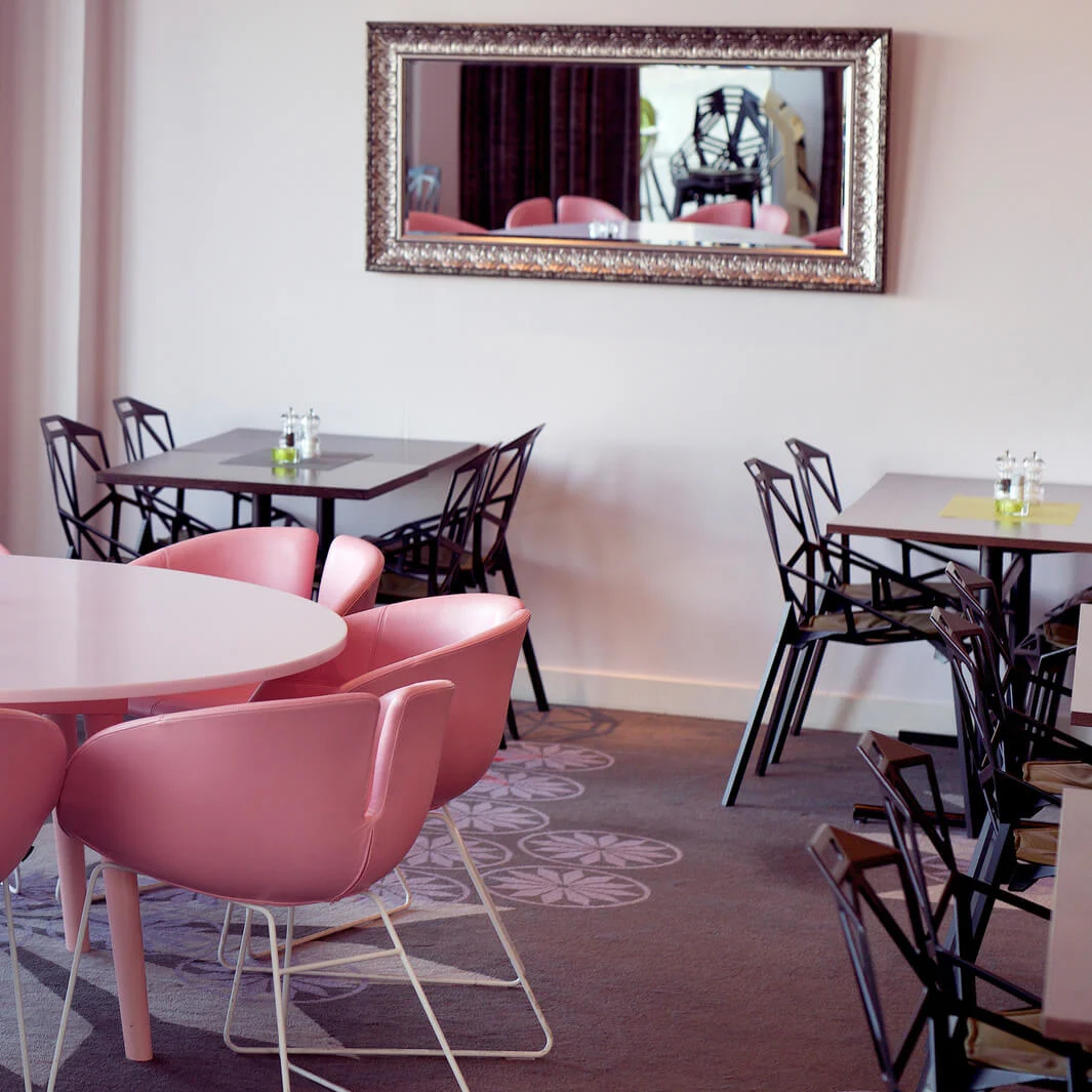 A round white table surrounded by pink chairs, inside an empty, brightly lit café with black tables, large windows, and a mirror on the wall.