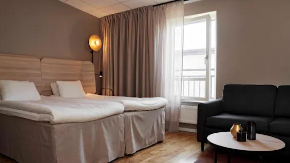 Ett bekvämt hotellrum med två sängar, en soffa och ett öppet fönster som släpper in naturligt ljus.