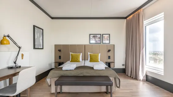A neatly arranged hotel room features a large bed with yellow cushions and folded towels, a desk with a yellow lamp, and artwork on the walls. Light fills the room through a window beside beige curtains.
