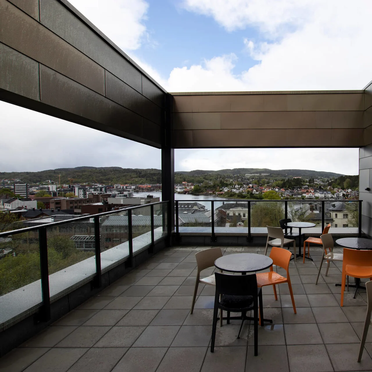 Borde og stole er placeret på en tagterrasse med udsigt over bylandskabet. Omgivelserne er udendørs og omgivet af glasrækværk, der indikerer et moderne bymiljø.