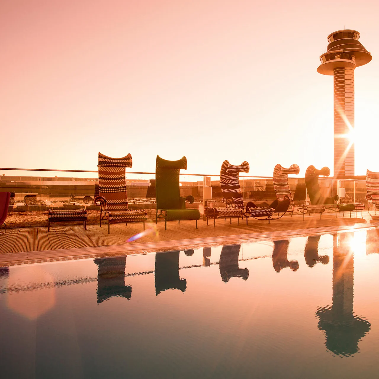 Poolside chairs face a reflecting pool at sunrise, beside a modern tower on a rooftop deck, with soft orange lighting.