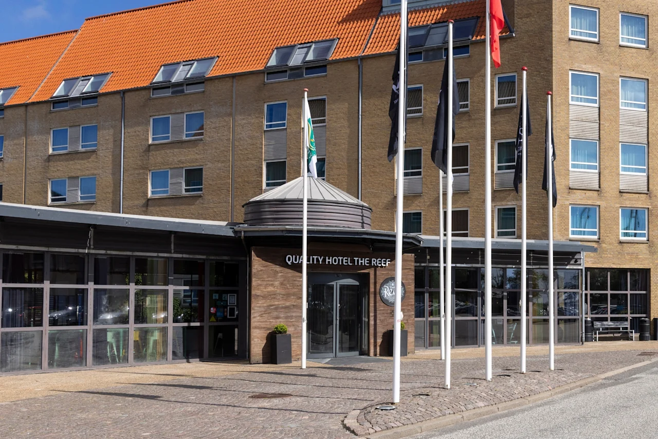 Hotellets indgang med skiltet "QUALITY HOTEL THE REEF" er omgivet af høje flagstænger. Bygningen har flere vinduer, et orange tegltag og en klar blå himmel i baggrunden.
