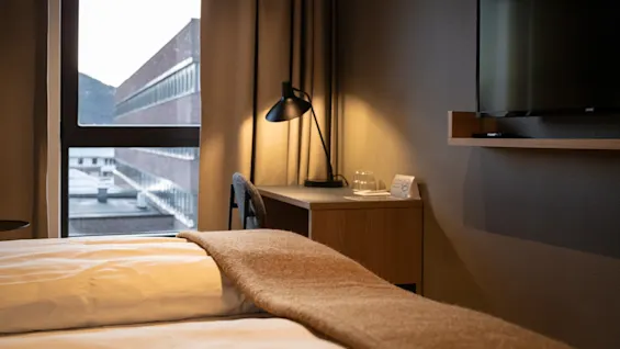 A comfortable hotel room featuring a bed with a blanket, a desk with a lamp, and a window overlooking a building.