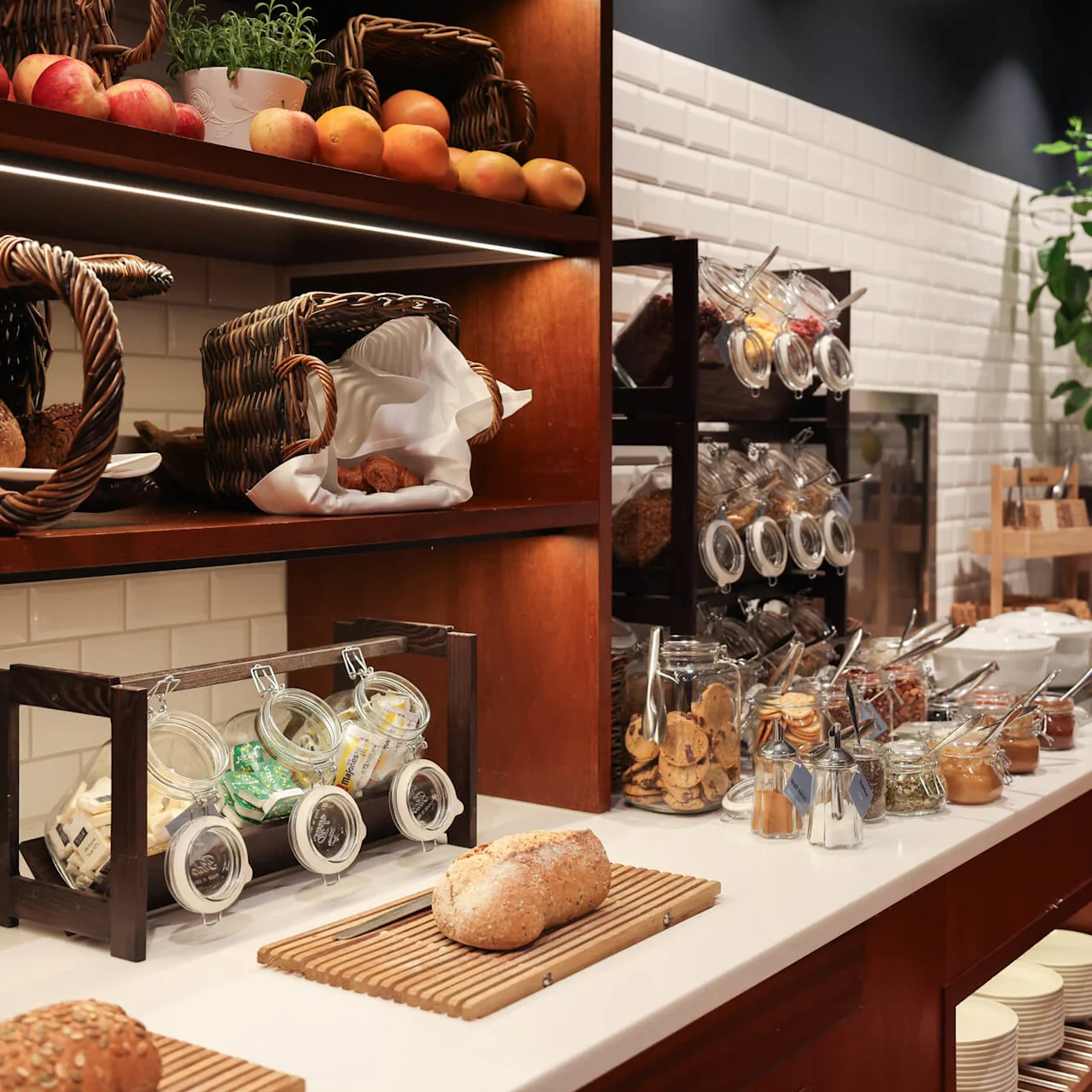 Bread loaves and baskets of fruits sit on a wooden shelf; jars filled with cereals and condiments line a white countertop. A potted plant adds greenery to the space.